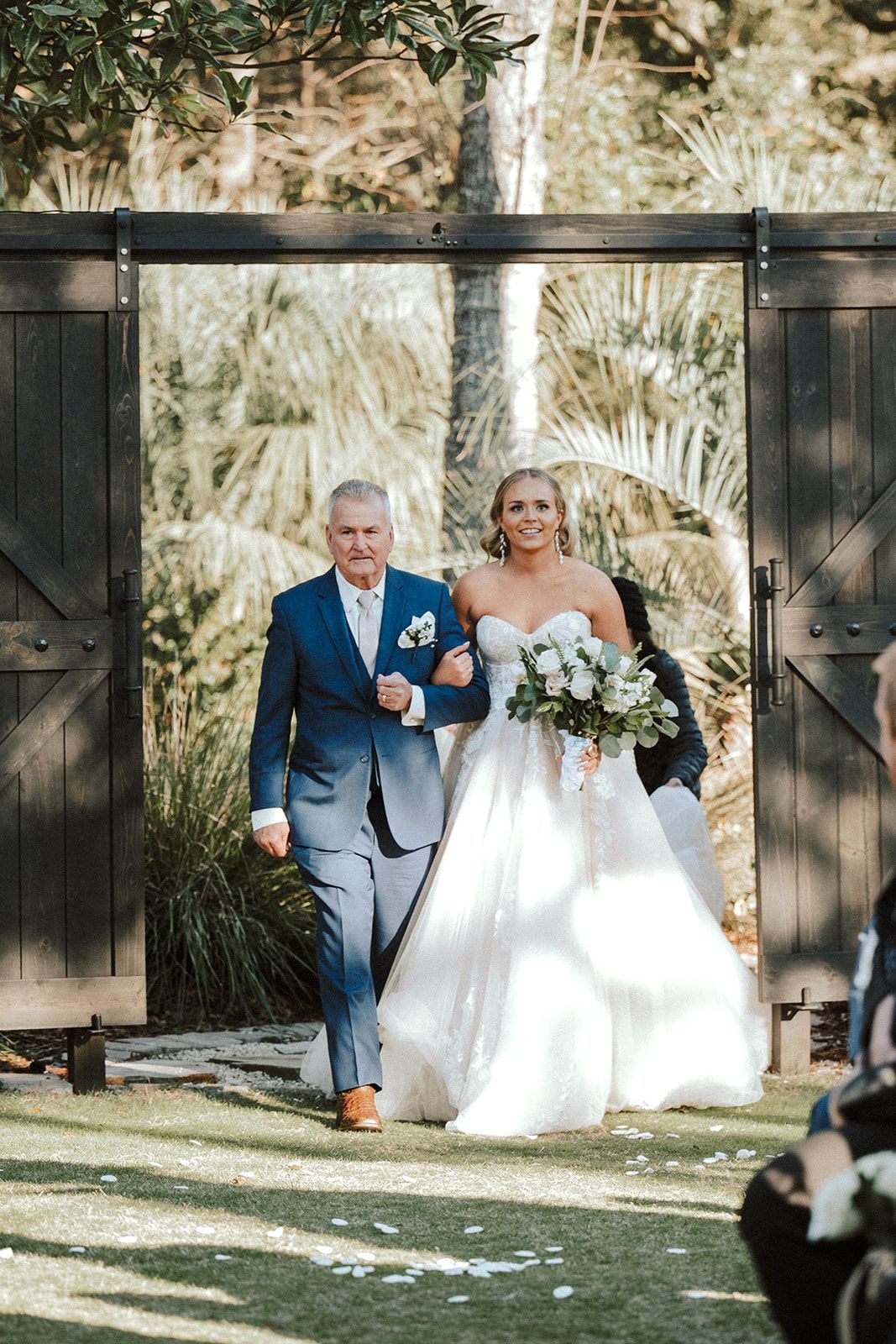 Bride and groom walking arm in arm through a barn doorway, bride in white gown holding bouquet, groom in blue suit