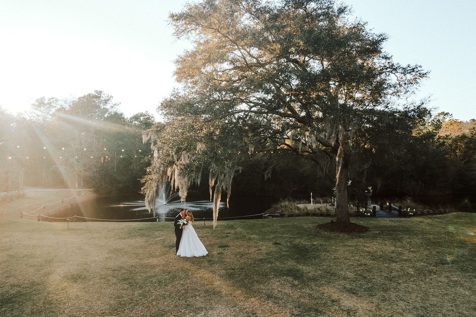Bride and groom stand in a sunlit grassy field beneath large trees.