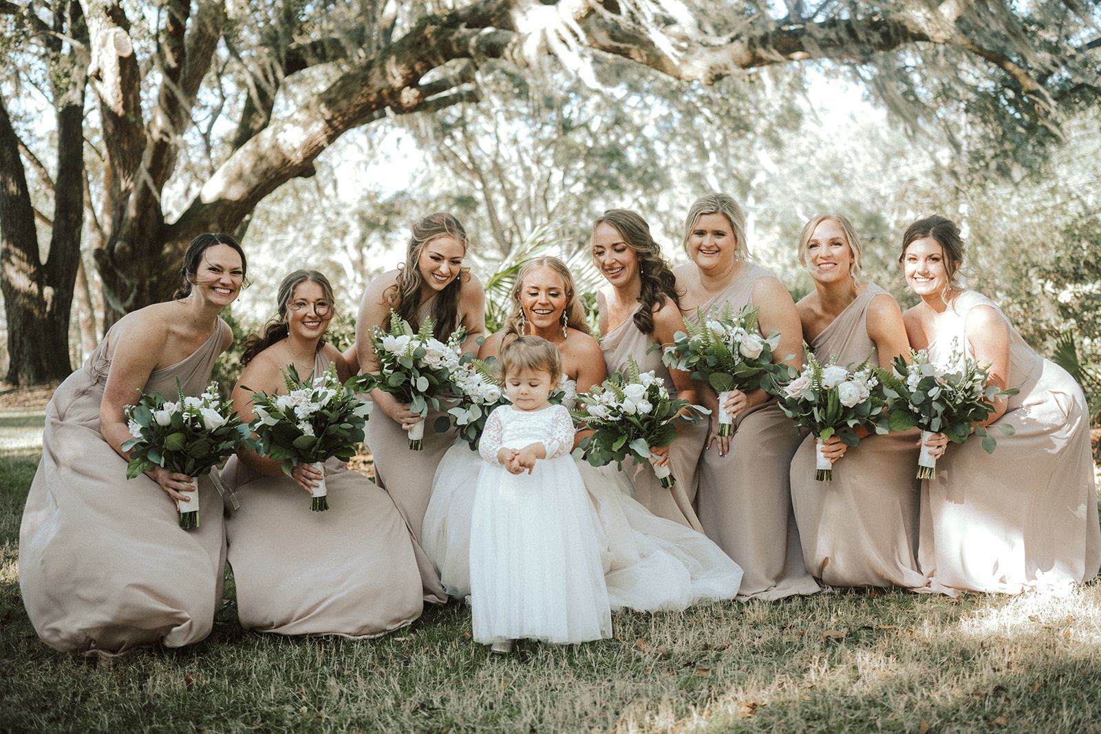 Bridal party posing outdoors with bouquets under a large tree, wearing blush and white dresses