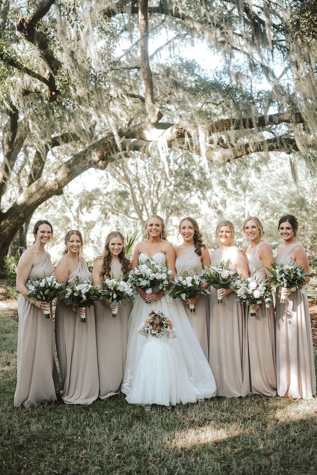 Bridal party in blush dresses standing outdoors under trees, holding white and greenery bouquets