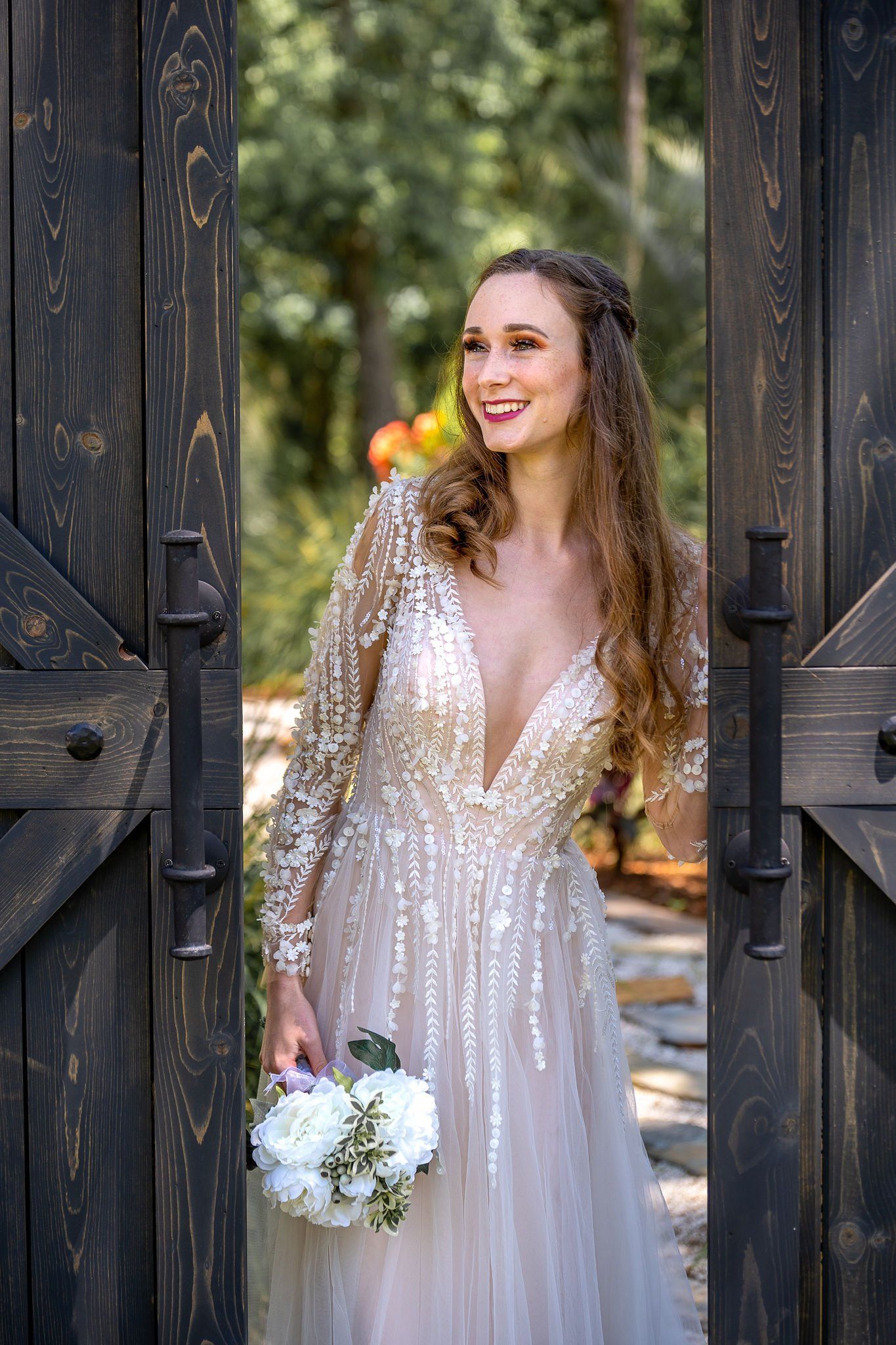 Bride in a sparkling white gown holding a bouquet, standing in a doorway and smiling outdoors.