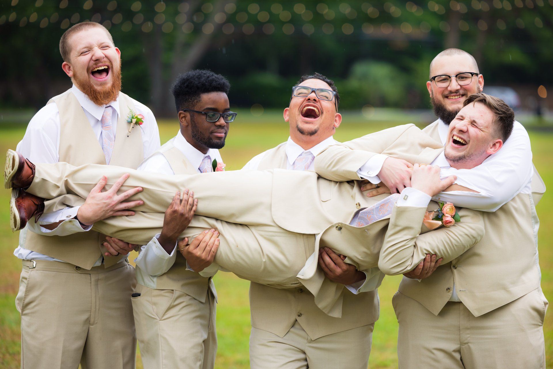 Five people in beige suits laughing while carrying a man outdoors on grass.