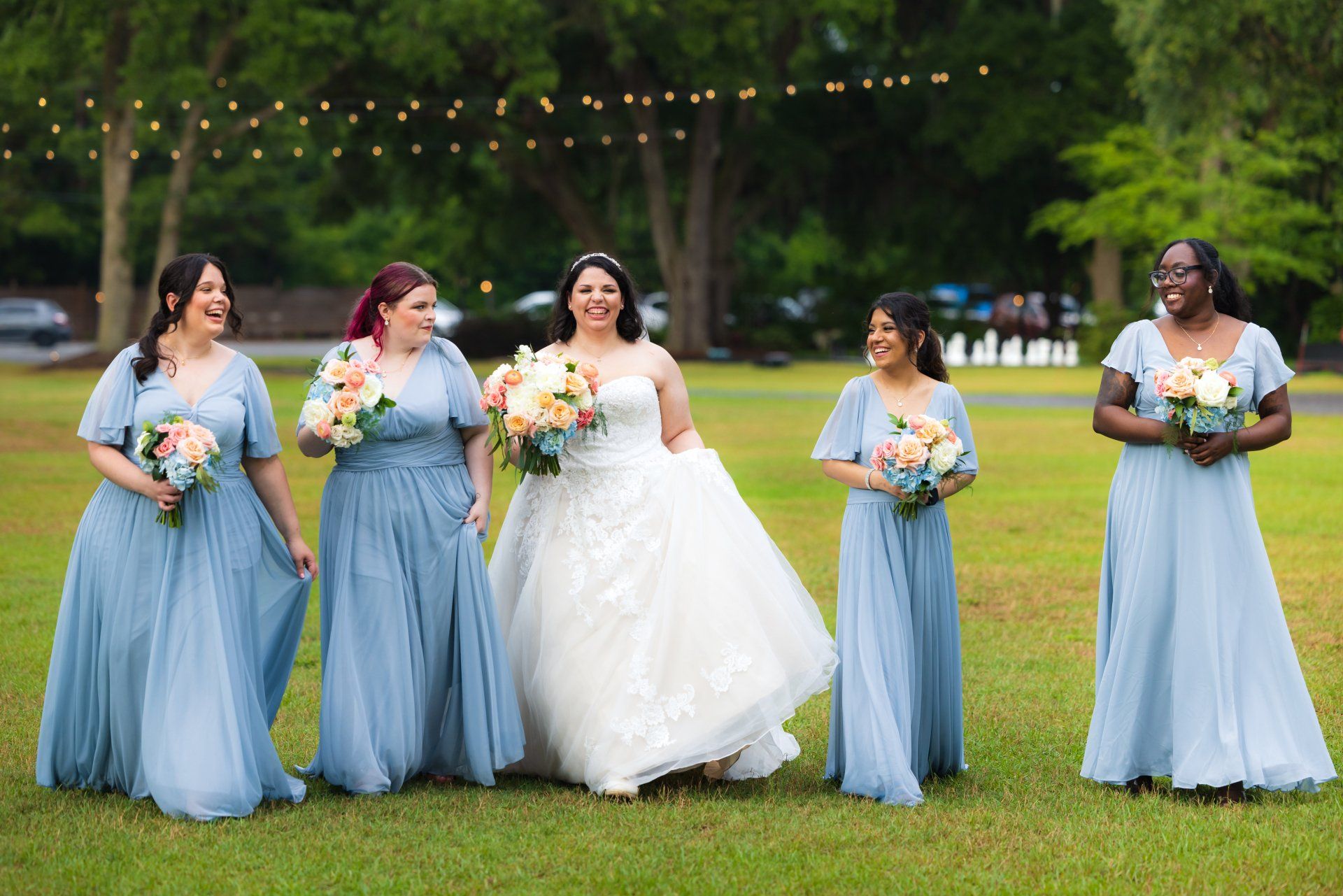 Five bridesmaids in light blue dresses and a bride in a white gown holding bouquets on a lawn