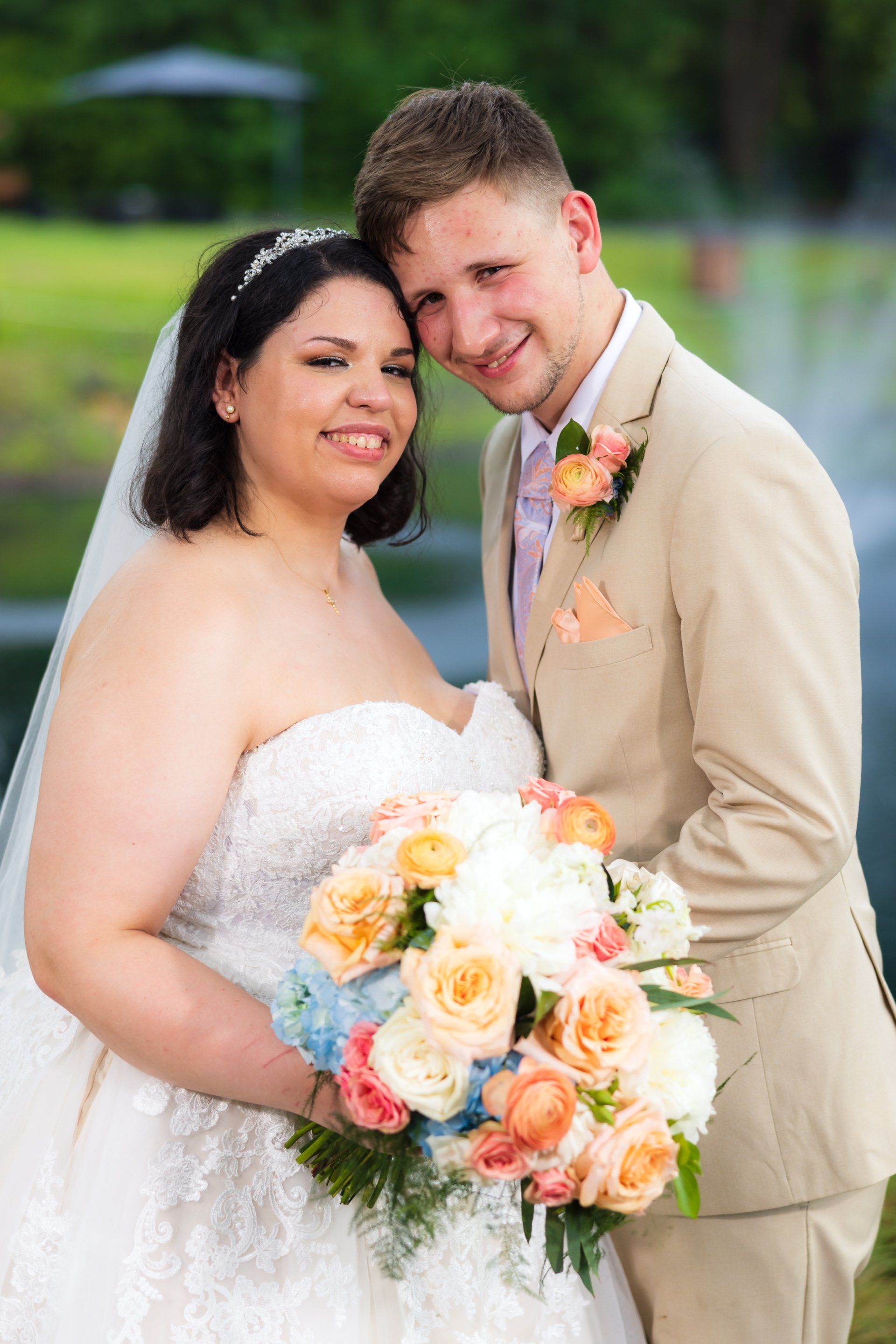 Bride and groom posing outdoors, smiling and holding a bouquet of peach and white flowers