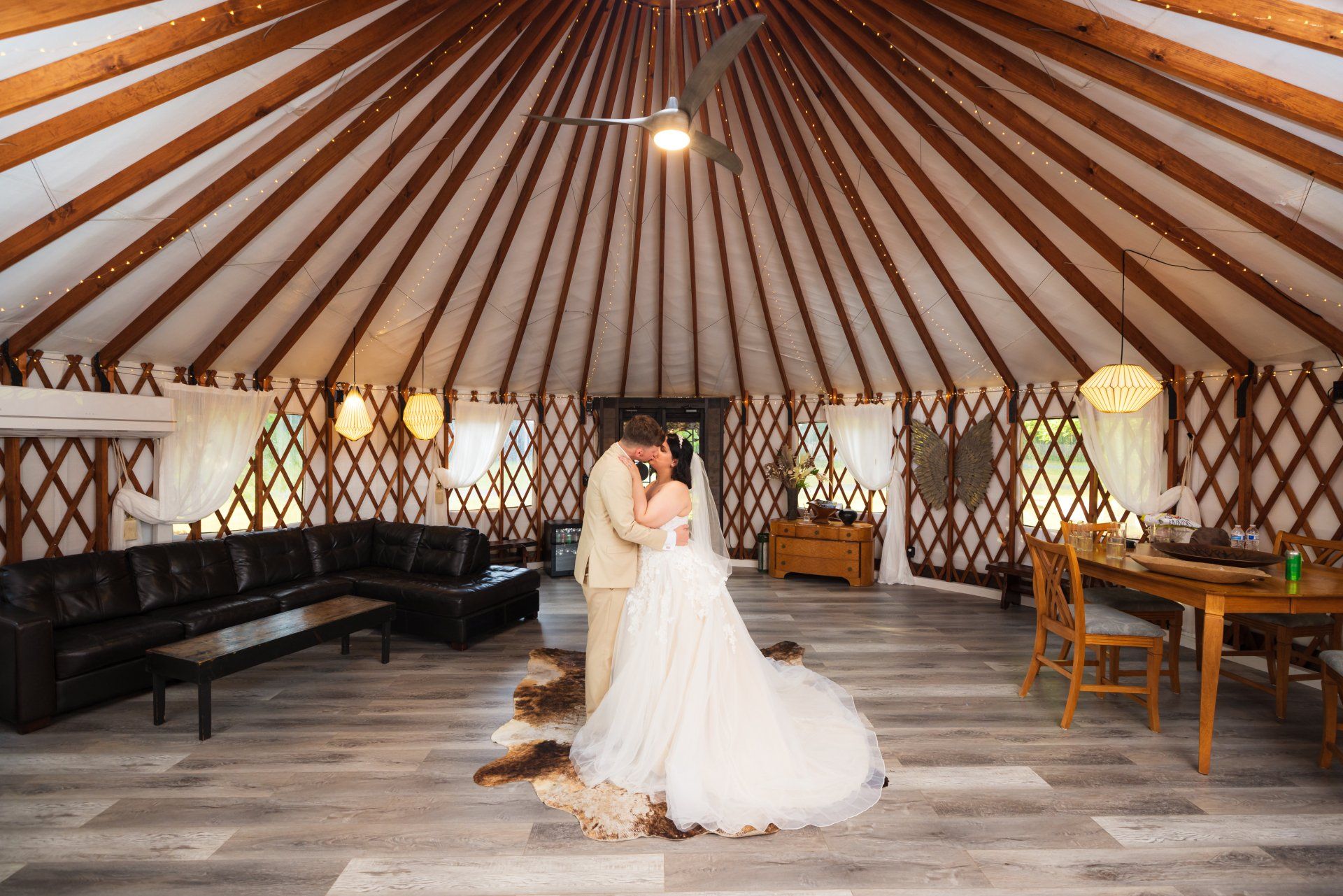 Bride in white gown standing in a yurt-style reception tent with rustic decor and wooden tables.