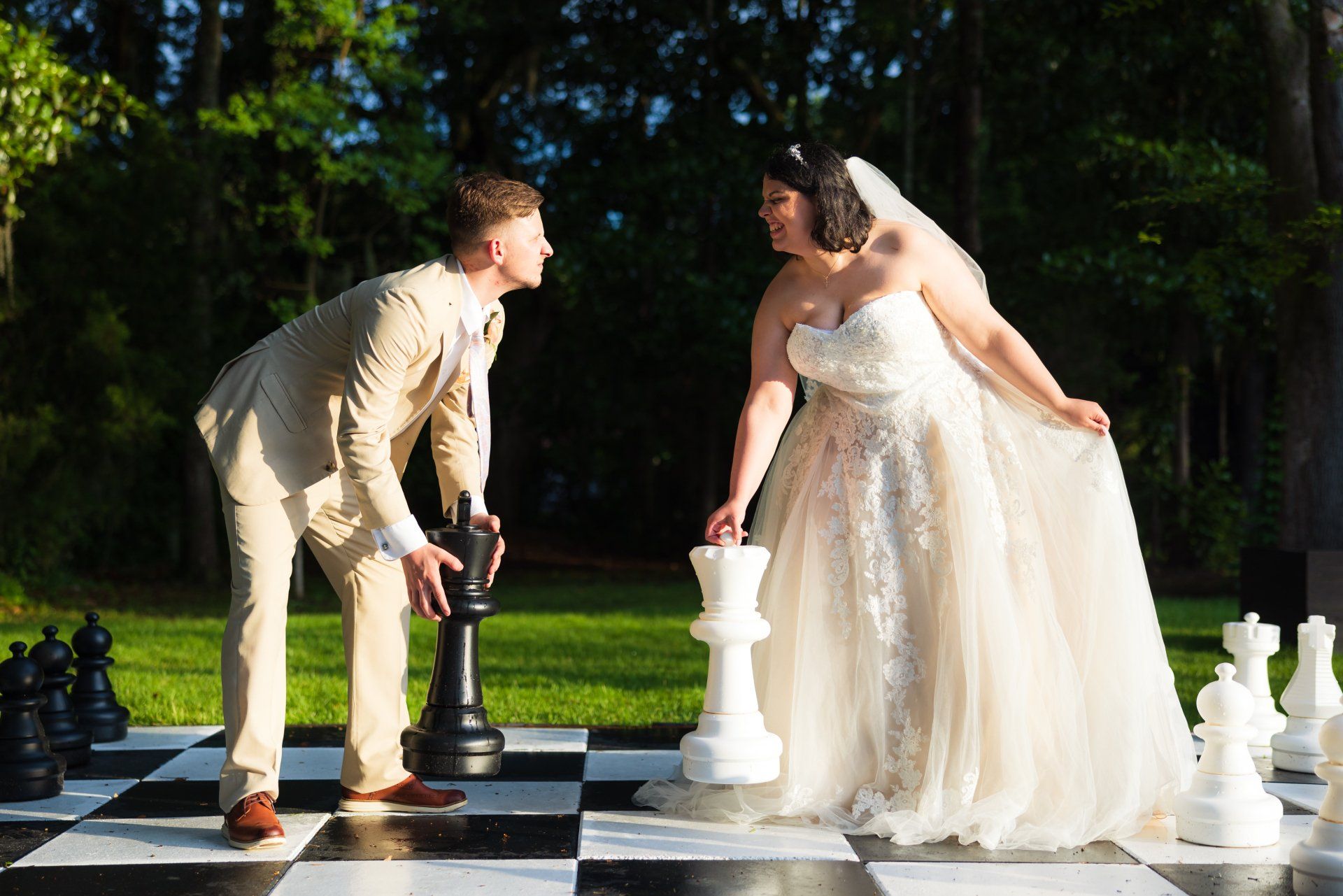 Bride and groom face off on a giant outdoor chessboard, leaning toward each other in wedding attire.