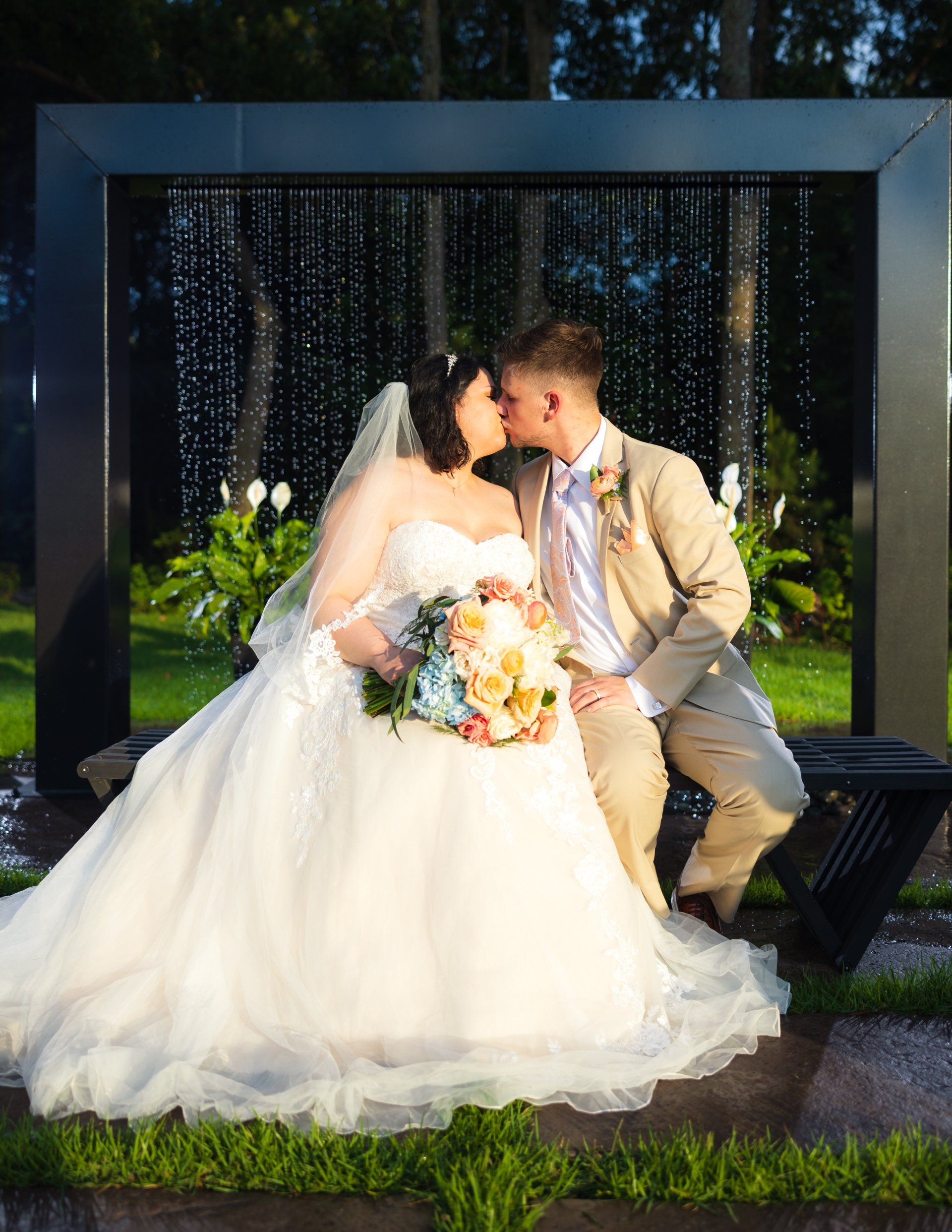 Wedding couple kissing on a bench under a black arch with hanging lights, bride in white gown holding bouquet