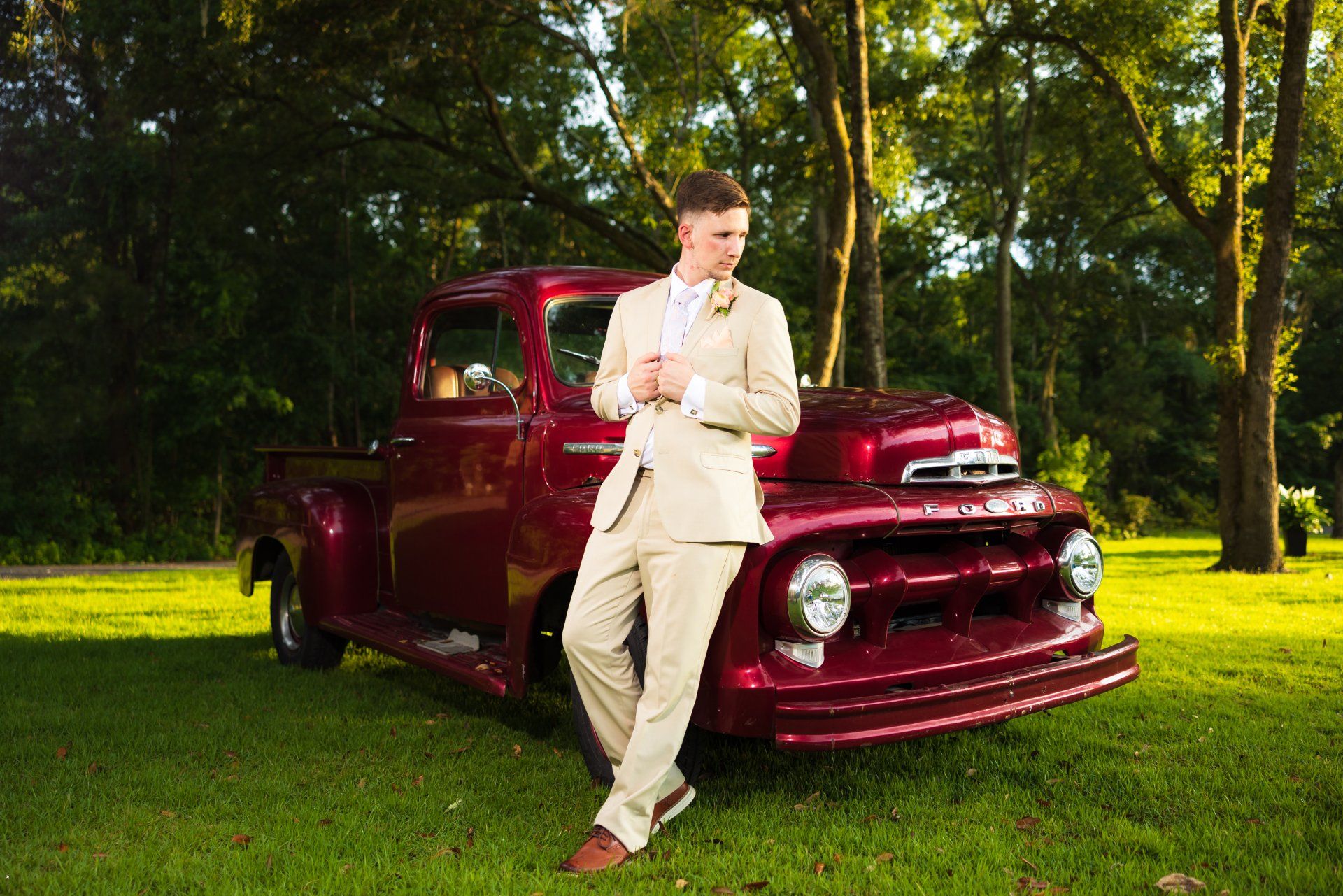 Man in beige suit leaning against a maroon vintage car on a grassy lawn under trees