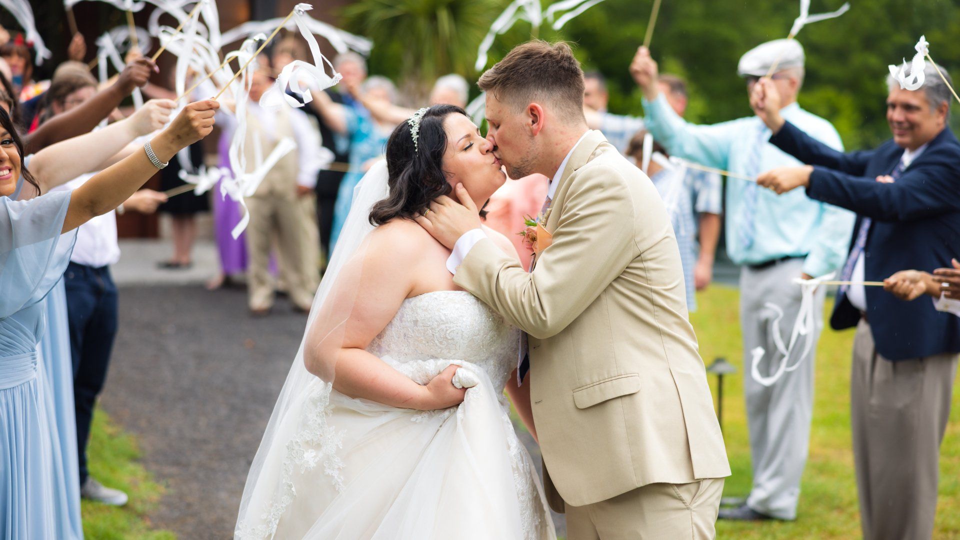 Bride and groom kiss under an arch of guests holding white ribbons at an outdoor wedding ceremony