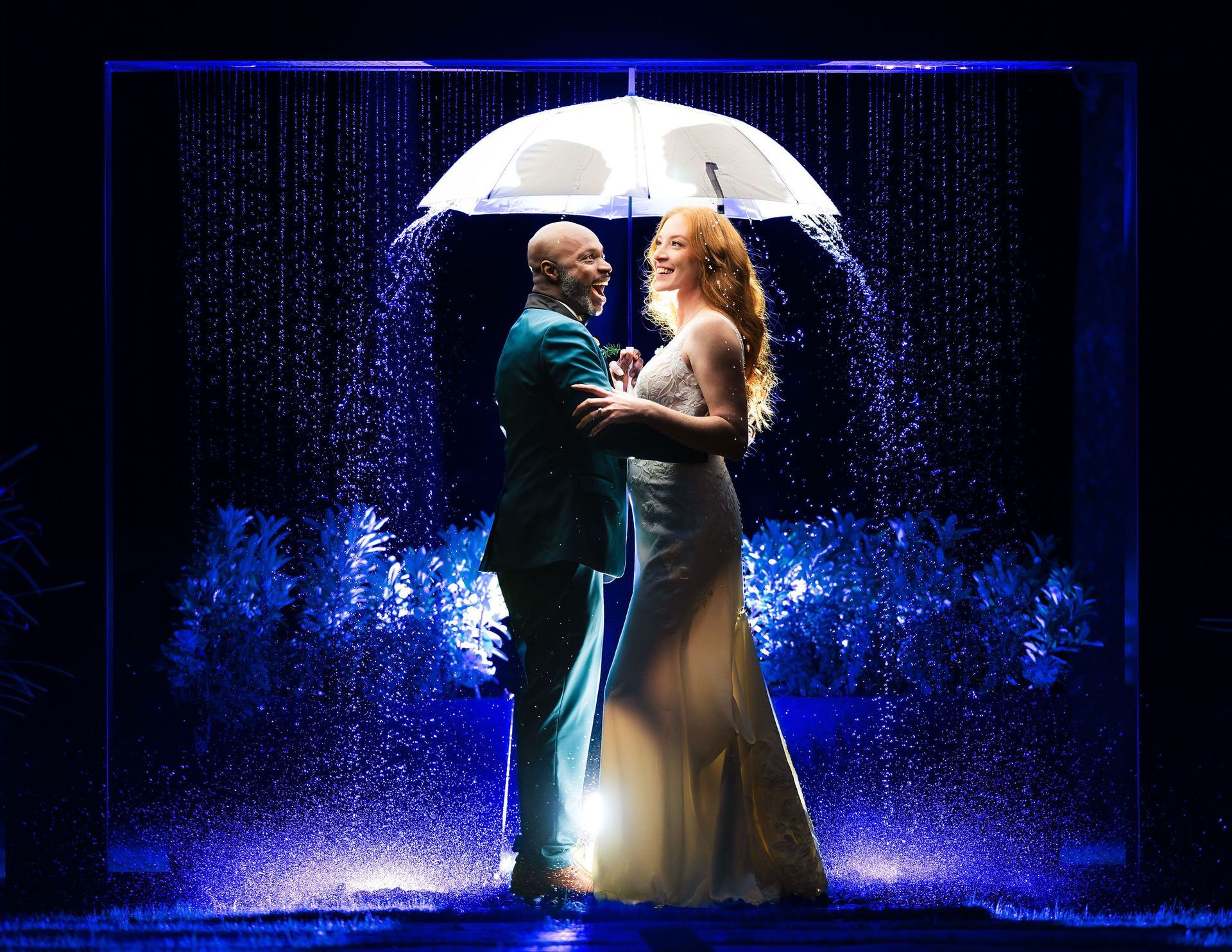 Two performers in formal attire embrace under a glowing umbrella amid blue stage lights and falling water