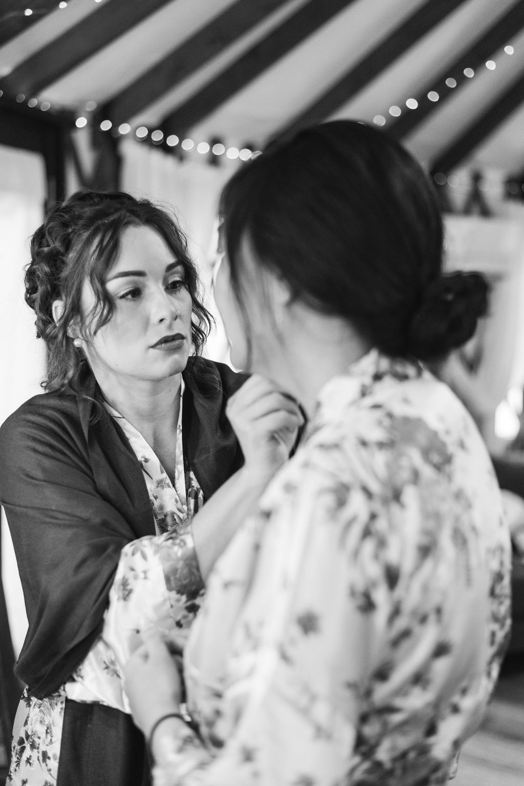 Two women talking indoors, one adjusting the other’s makeup in a softly lit setting.