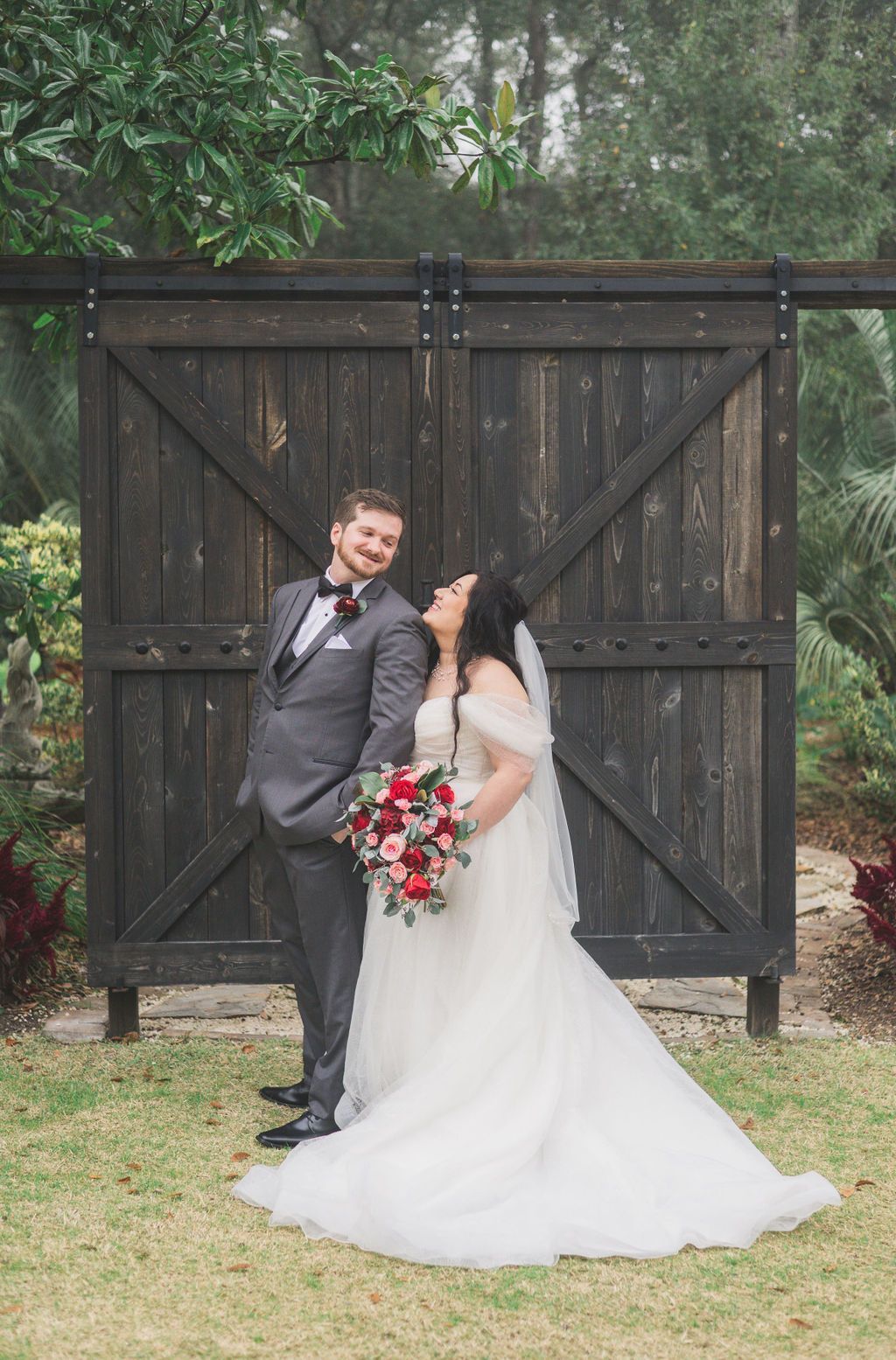 Bride and groom posing in front of a large wooden gate, bride holding a red and white bouquet.