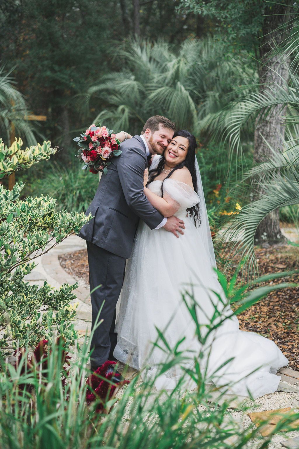 Bride and groom kissing in a lush garden, bride holding a bouquet of red and white flowers.