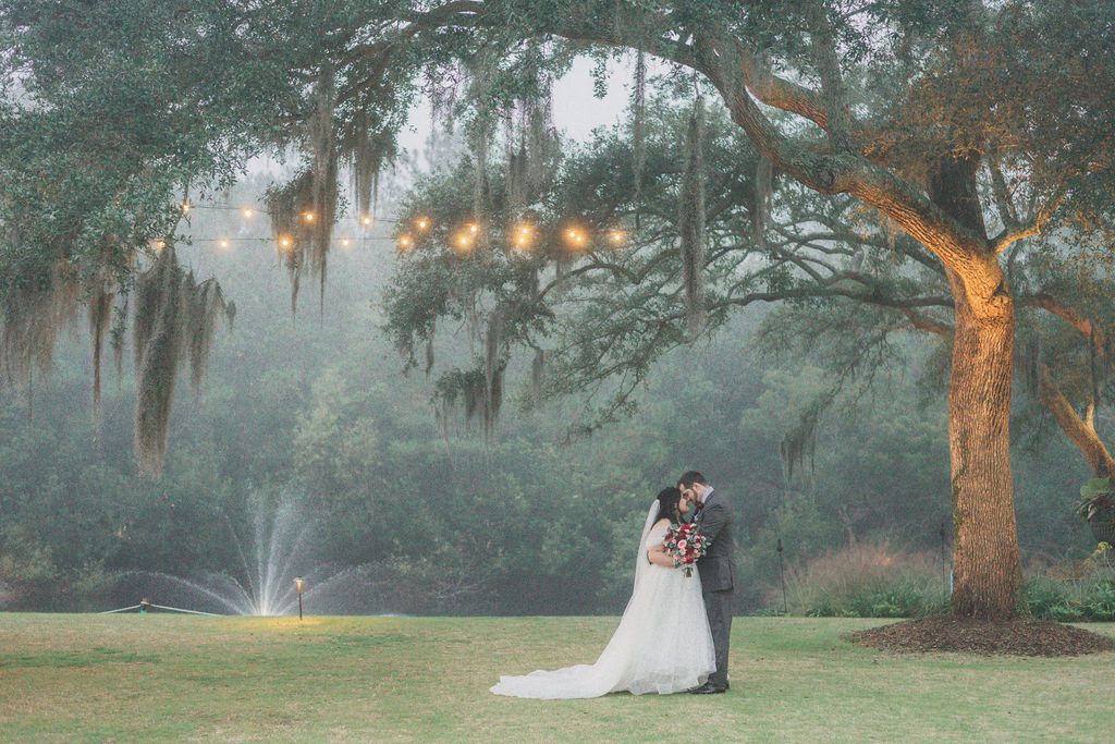 Bride and groom kissing under a large tree in a misty garden with fountain lights