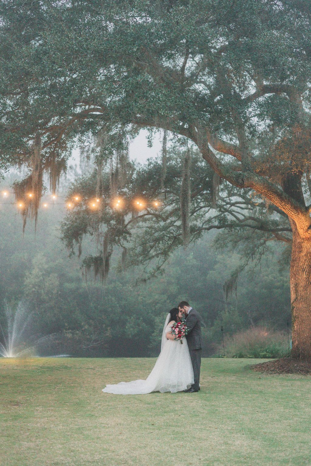 Couple in wedding attire embracing under a large tree at dusk, with warm lights and misty greenery.