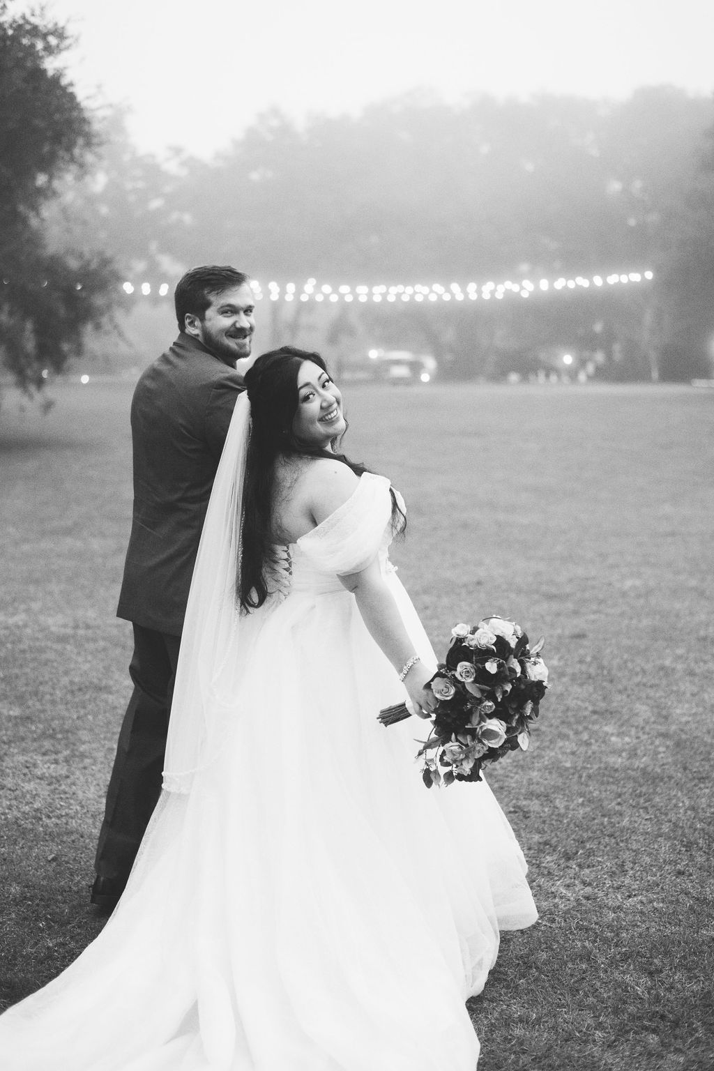 Bride and groom posing on a foggy lawn, bride holding a bouquet and smiling over her shoulder