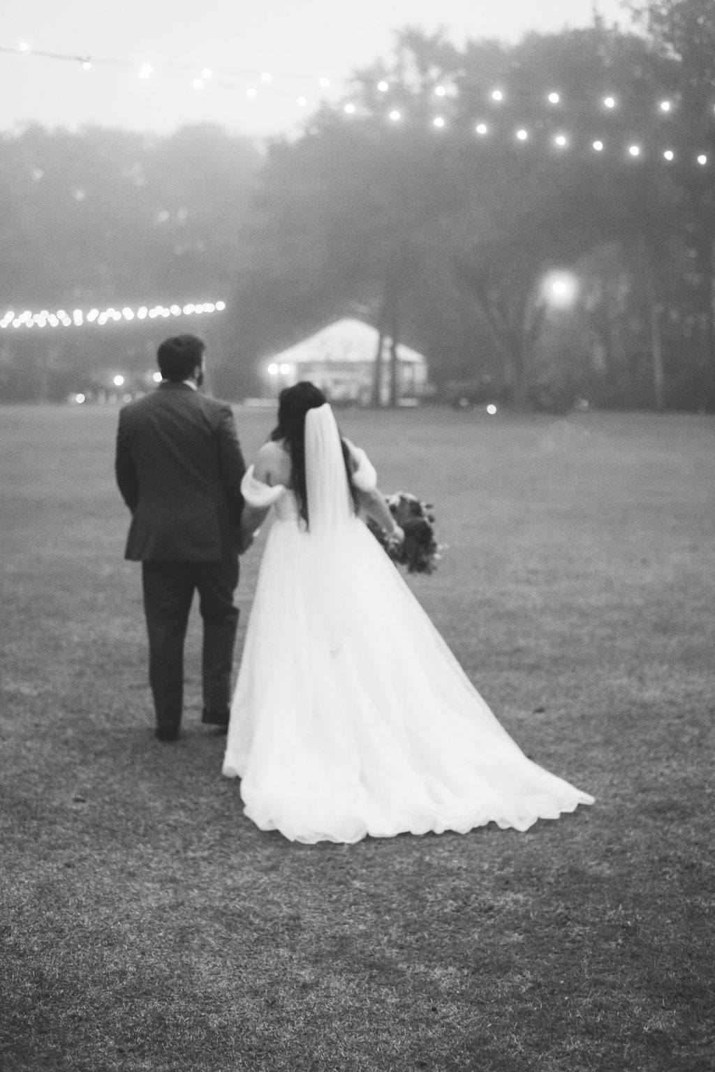 Bride and groom walking across a grassy field at dusk under string lights