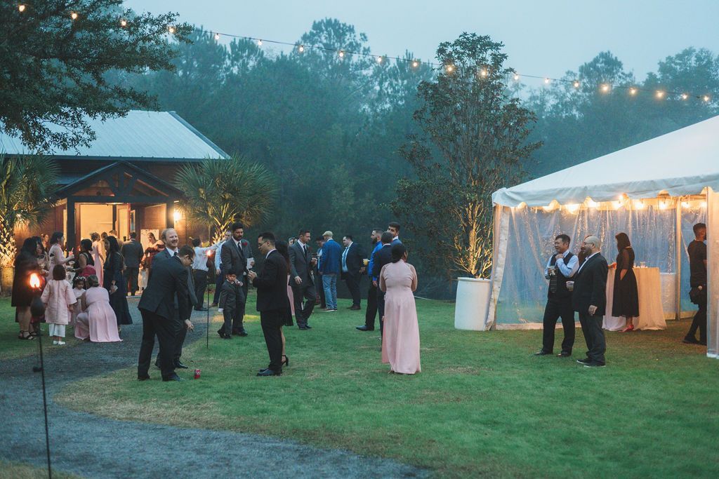 Outdoor evening wedding reception with guests mingling beside a lit tent and pavilion on a grassy lawn