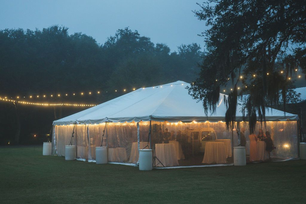 Illuminated white event tent at dusk on a grassy lawn, with string lights and trees in the background