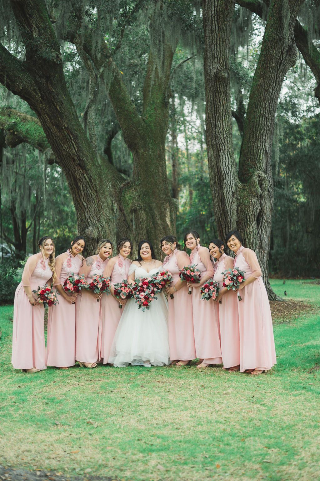 Seven bridesmaids and a bride in blush dresses stand under a large tree holding bouquets.
