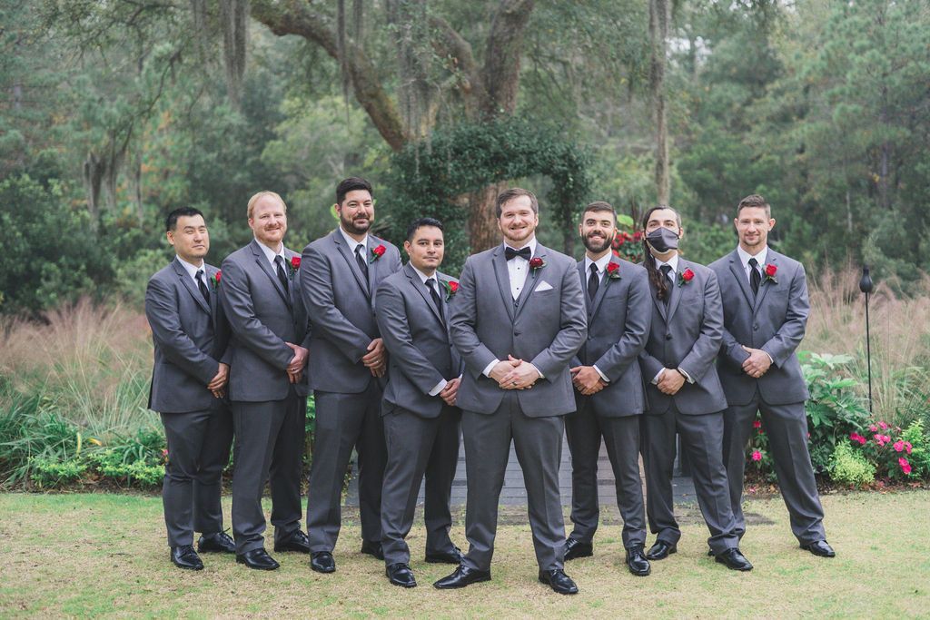 Nine men in dark suits and red ties stand outdoors in a wooded park.