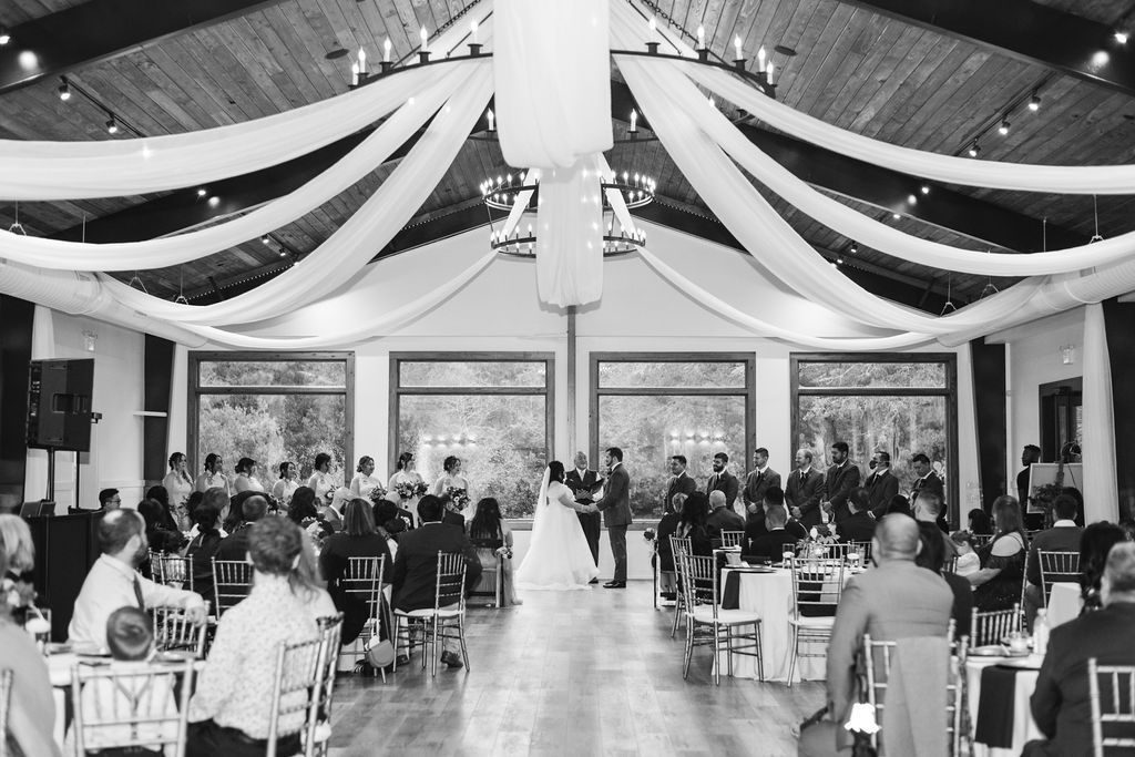 Indoor wedding reception with guests seated at tables beneath draped ceiling fabric, facing the couple at the front.