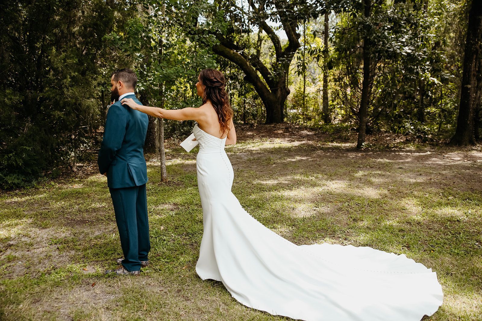 Bride in a white gown adjusting the collar of a man in a teal suit outdoors under trees