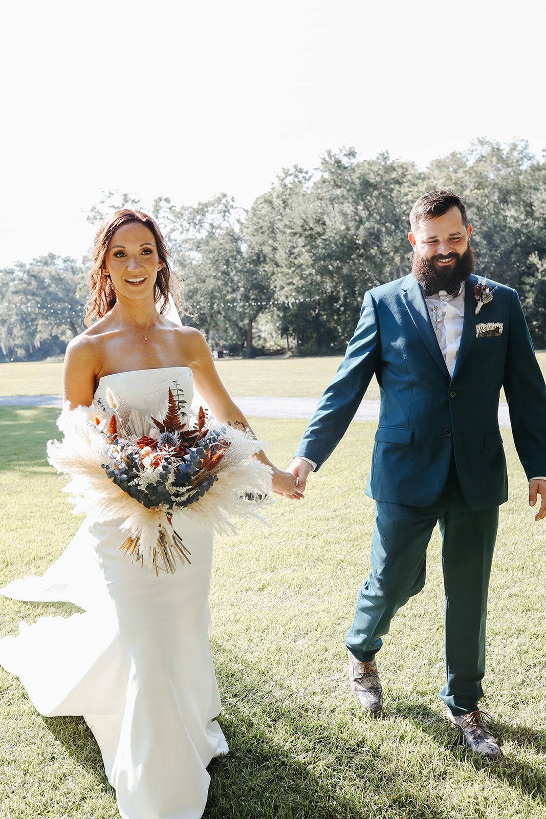 Bride and groom walking outdoors by a lake, holding hands, with bouquet and blue suit