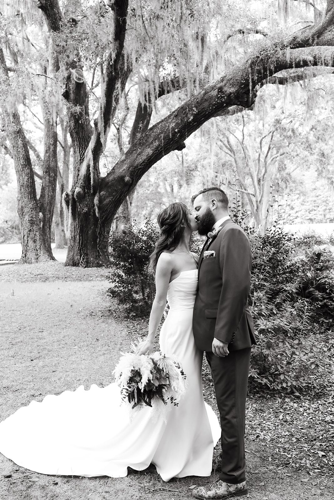 Bride and groom standing together outdoors beneath large trees, the bride holding a bouquet.