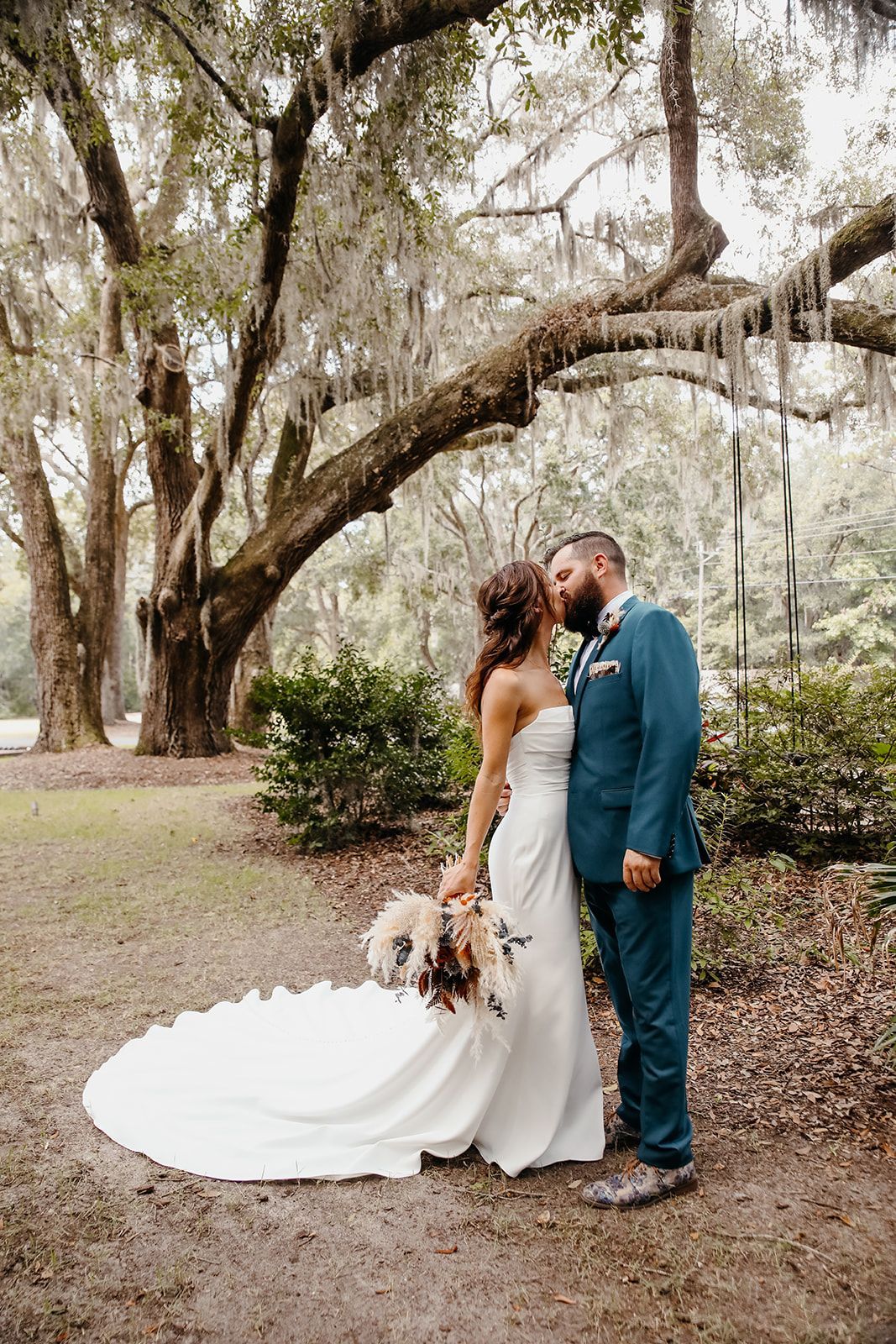 Bride and groom kiss beneath mossy trees, with a small dog at the bride’s side.