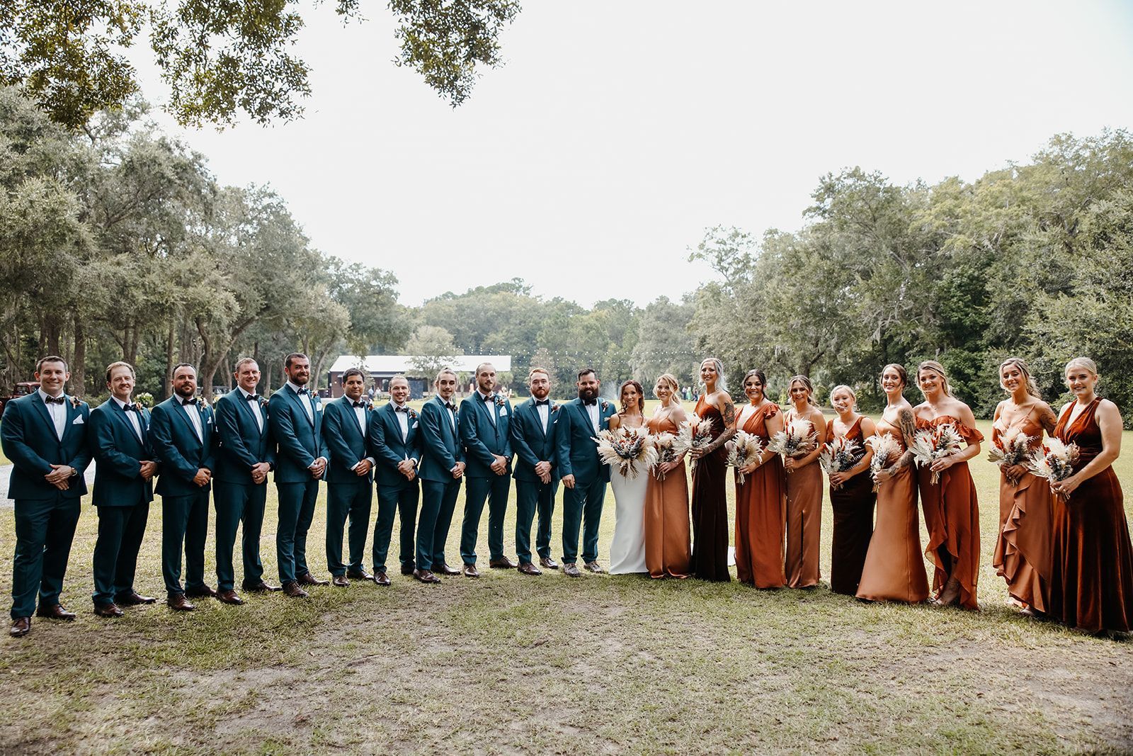 Wedding party posing outdoors in formal attire, with navy suits on one side and rust dresses on the other.