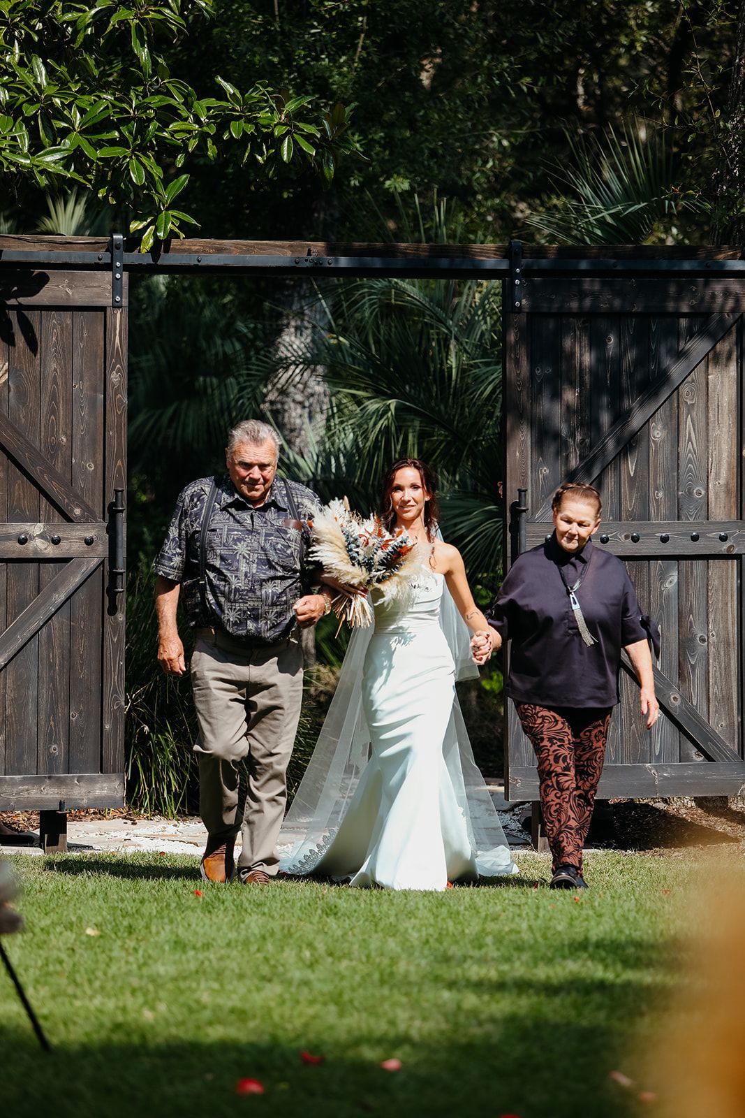 Bride walking with two men through large wooden gates in a garden setting