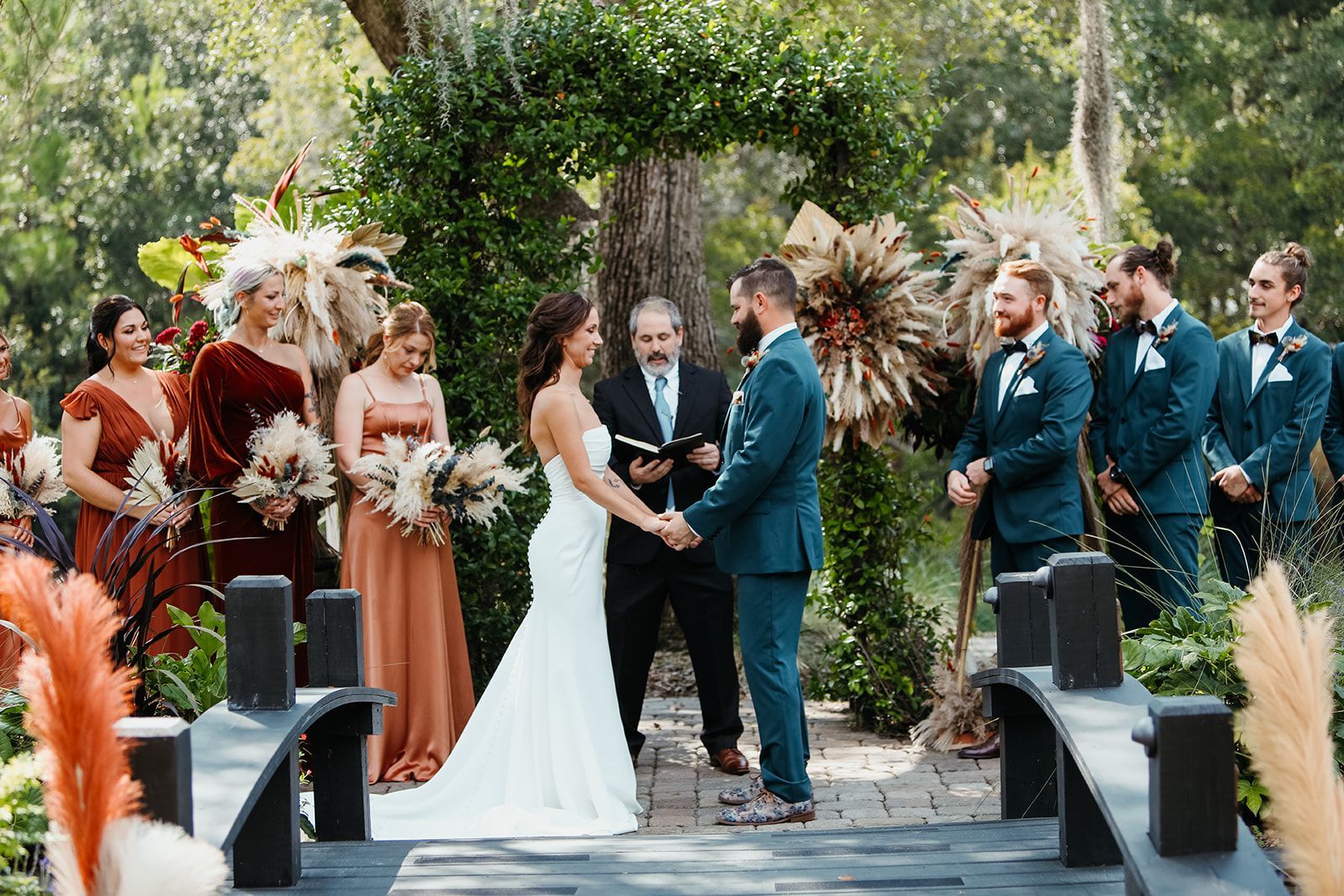 Wedding couple exchanging vows outdoors, surrounded by attendants in teal and rust attire amid greenery.