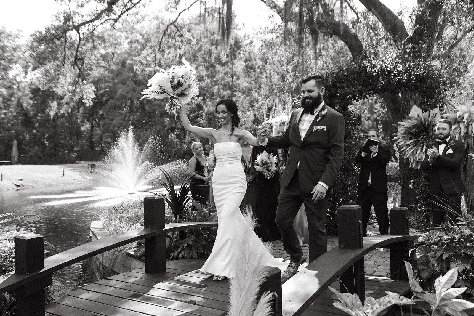 Newlyweds walking across a garden bridge beside a fountain, with guests behind them