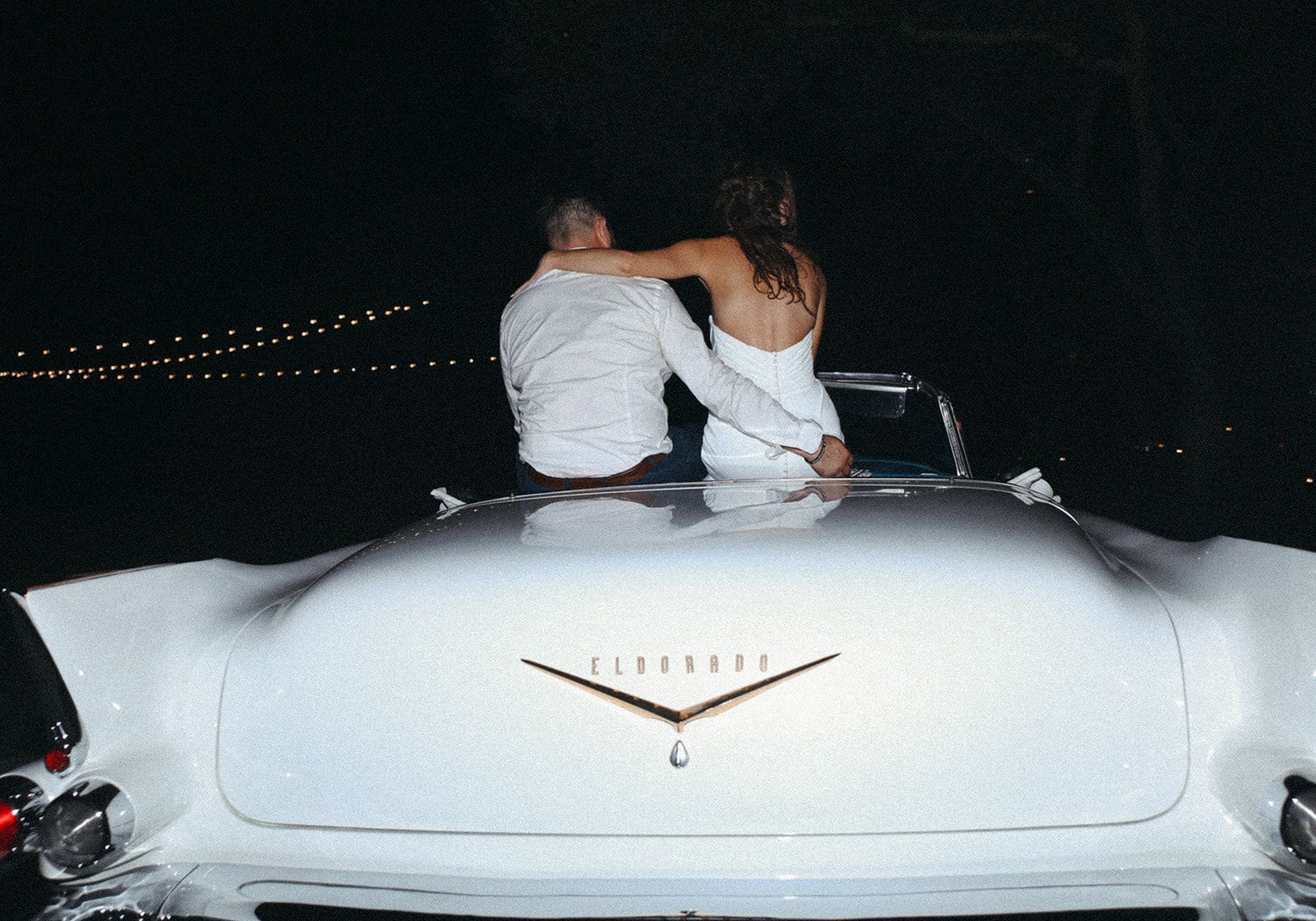 Two people sitting on the back of a white car at night, with city lights in the background
