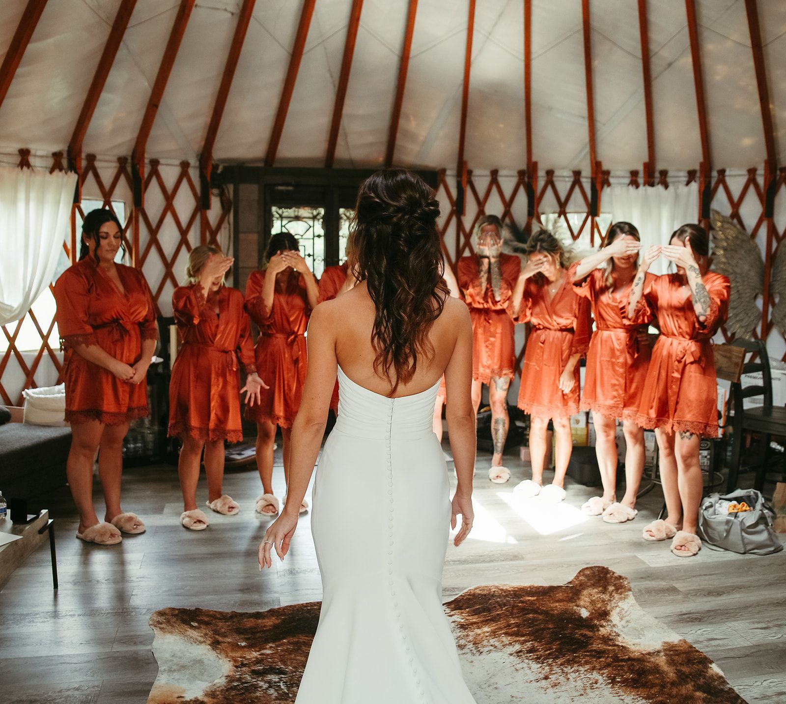 Woman in white dress facing a group in orange dresses inside a round ceremonial tent