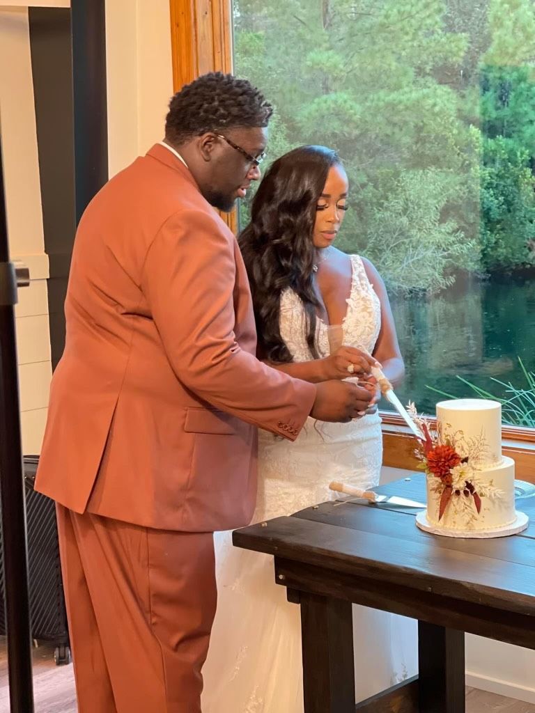 Two people cut a white wedding cake at a table by a large window.
