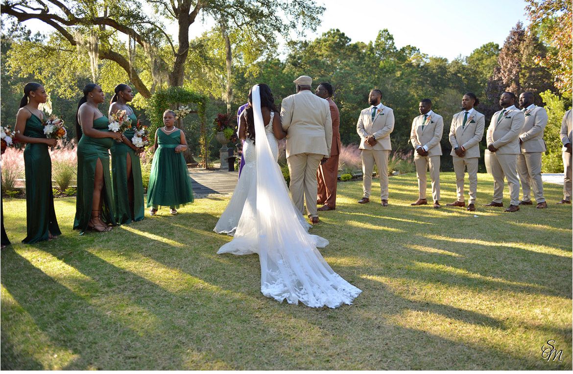 Wedding party standing on a grassy lawn as bride walks toward groom during outdoor ceremony