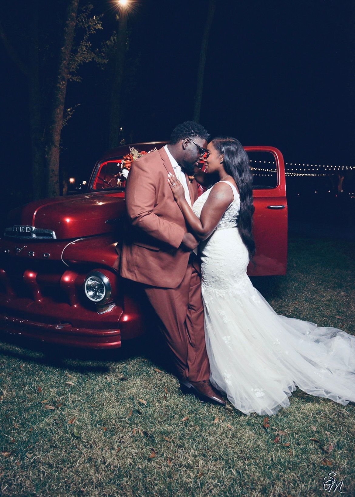 Couple kissing beside a red vintage car at night, bride in white gown and groom in tan suit