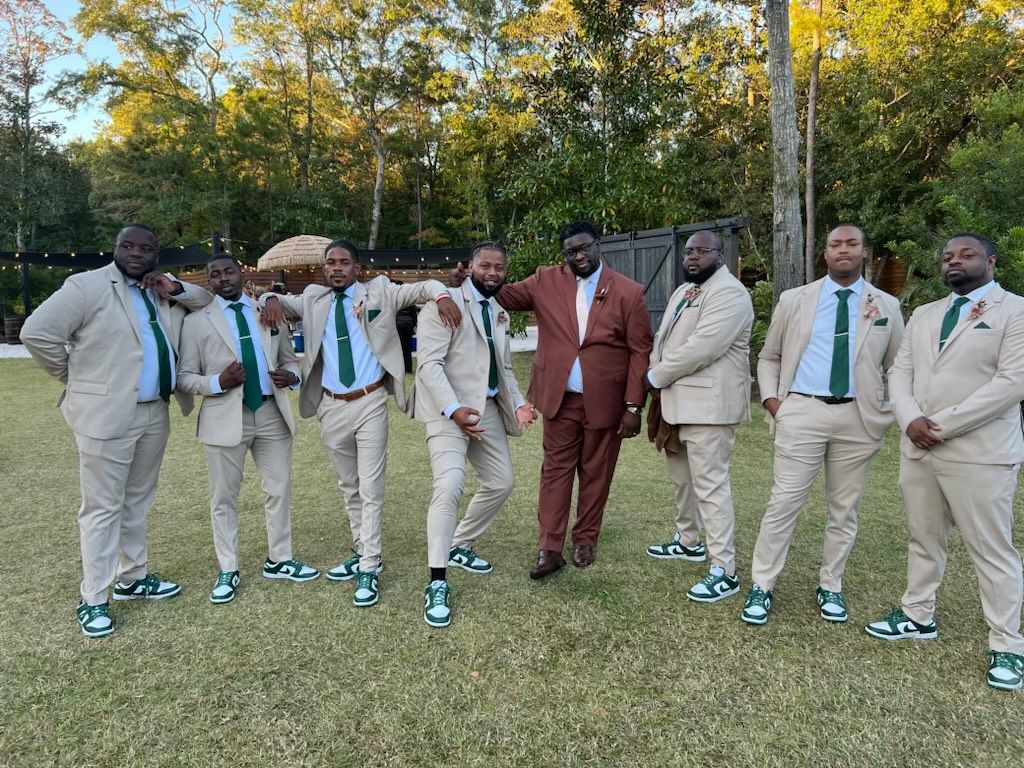 Eight men in coordinated beige suits pose together on a grassy path in a wooded park.