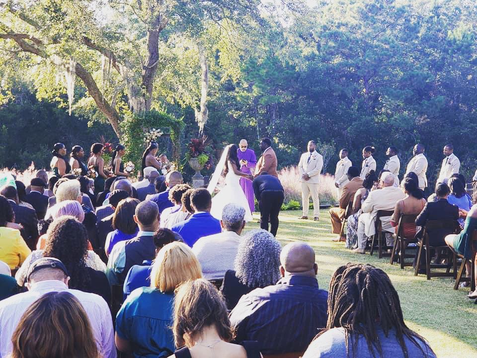 Outdoor wedding ceremony under trees, with guests seated on chairs and the couple standing at the front.