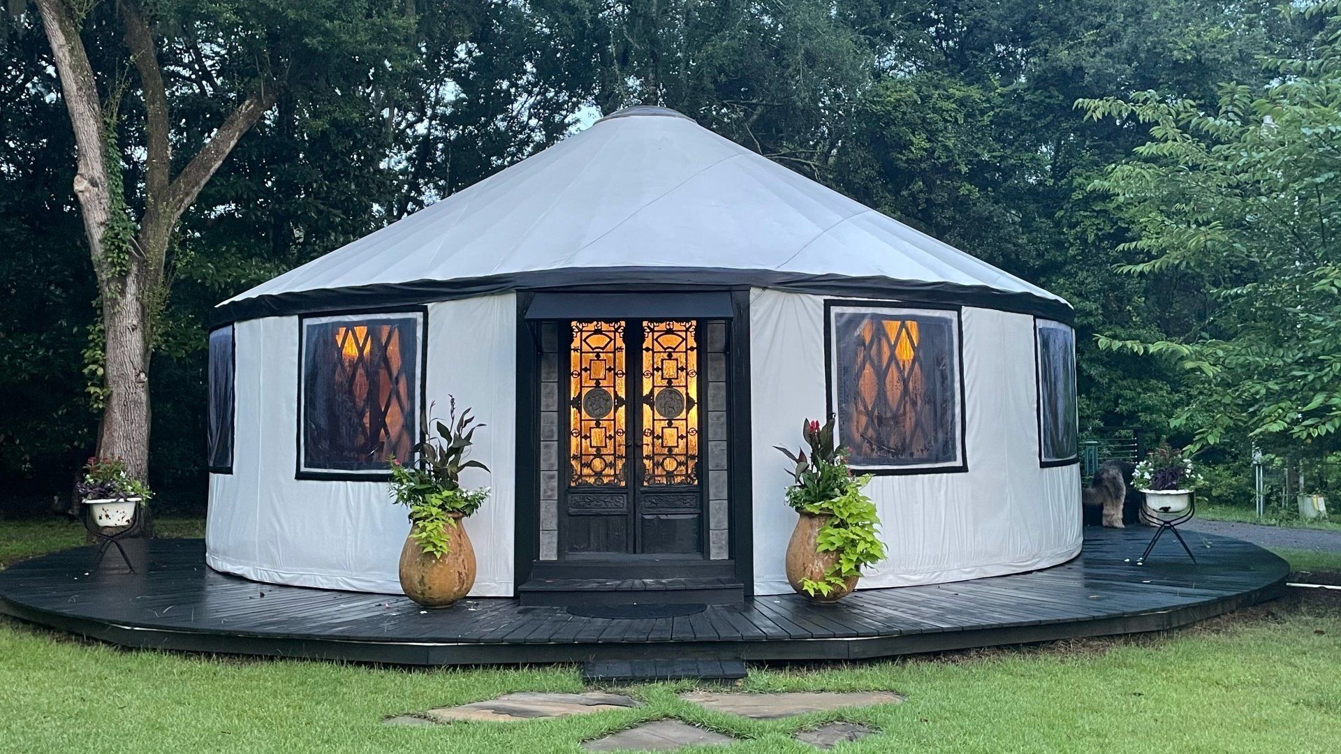Round white cottage with black roof and lit windows, surrounded by trees and greenery.