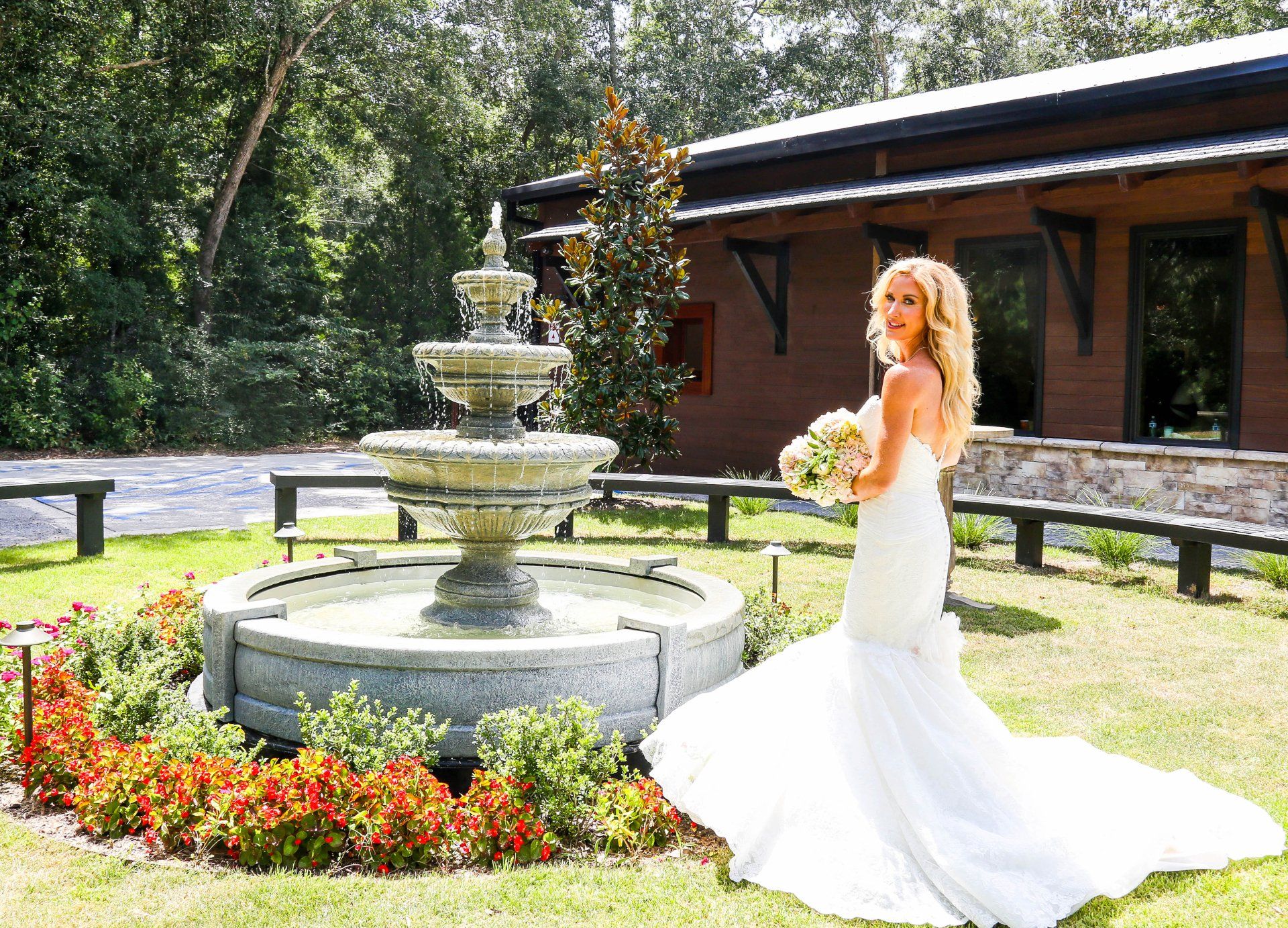 Bride in a white gown holding a bouquet beside a fountain in a sunny garden