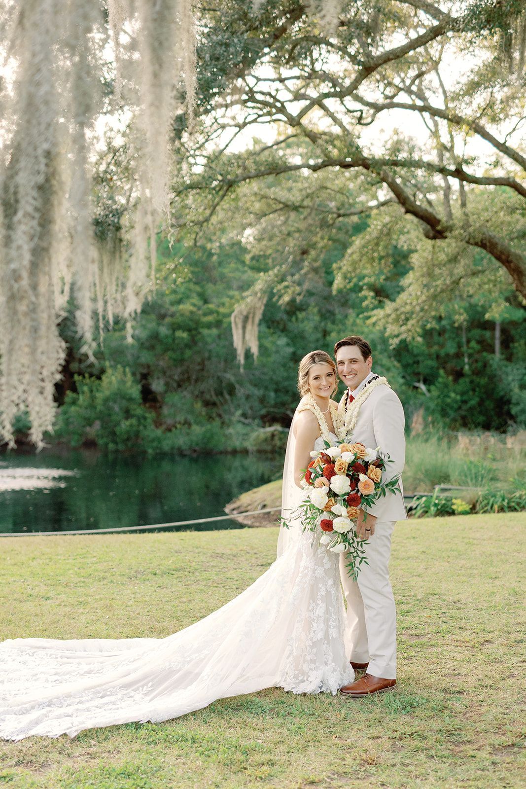 Bride and groom pose by a lake under hanging moss, with a long white gown and colorful bouquet.
