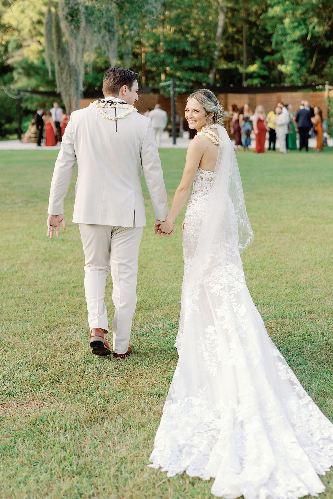 Bride and groom holding hands on a grassy lawn, walking away in wedding attire outdoors.
