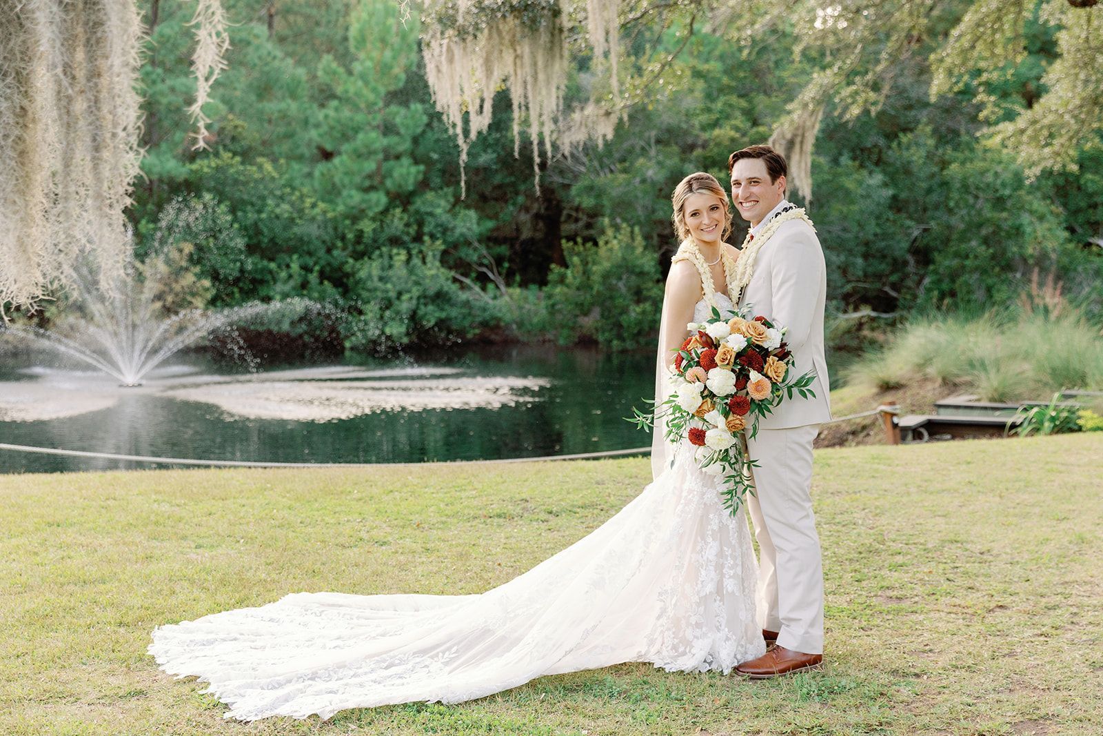 Wedding couple posing by a pond with a fountain, bride in white gown and groom in tan suit