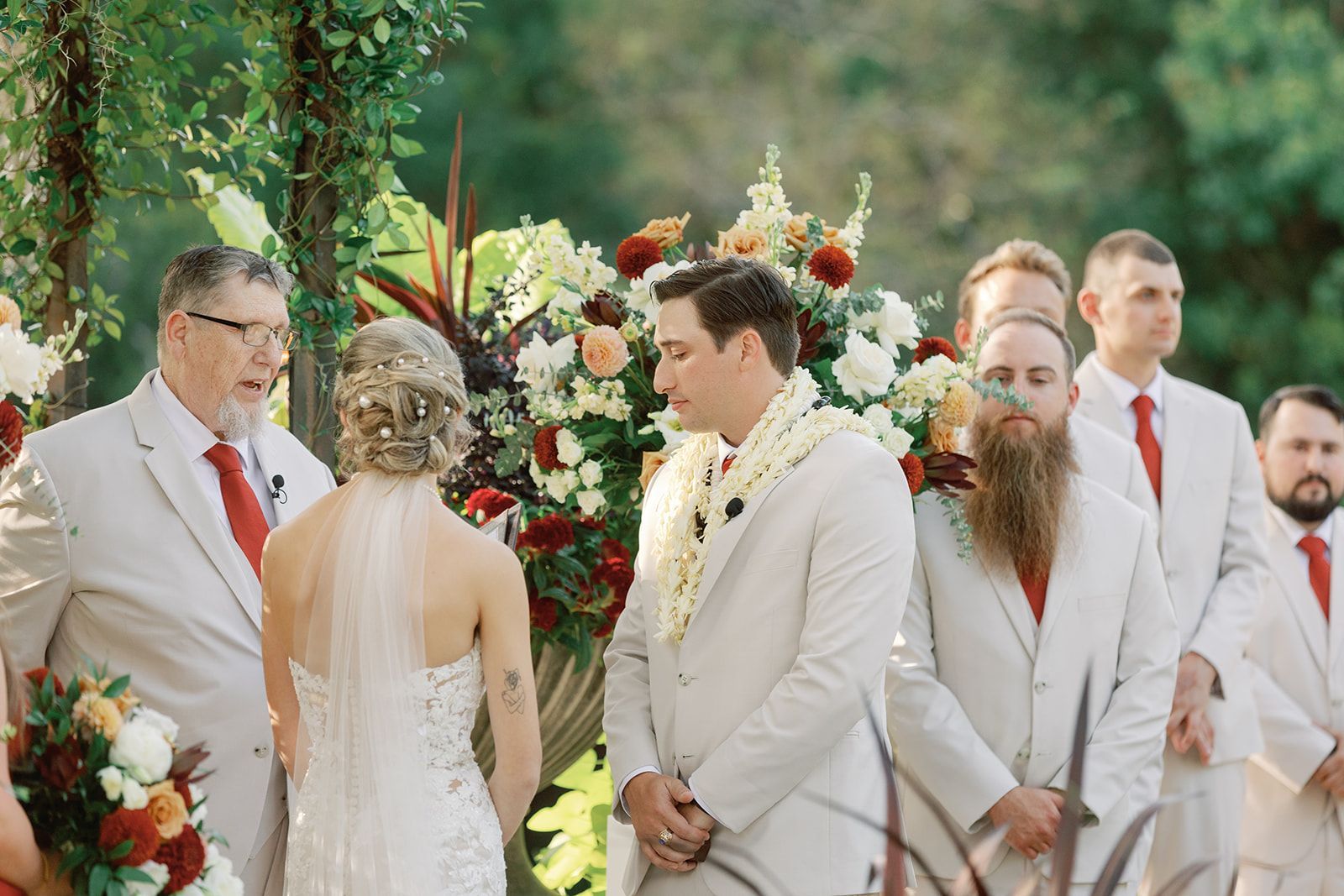 Wedding party gathered outdoors around floral arrangements, with the bride facing the groom and attendants nearby