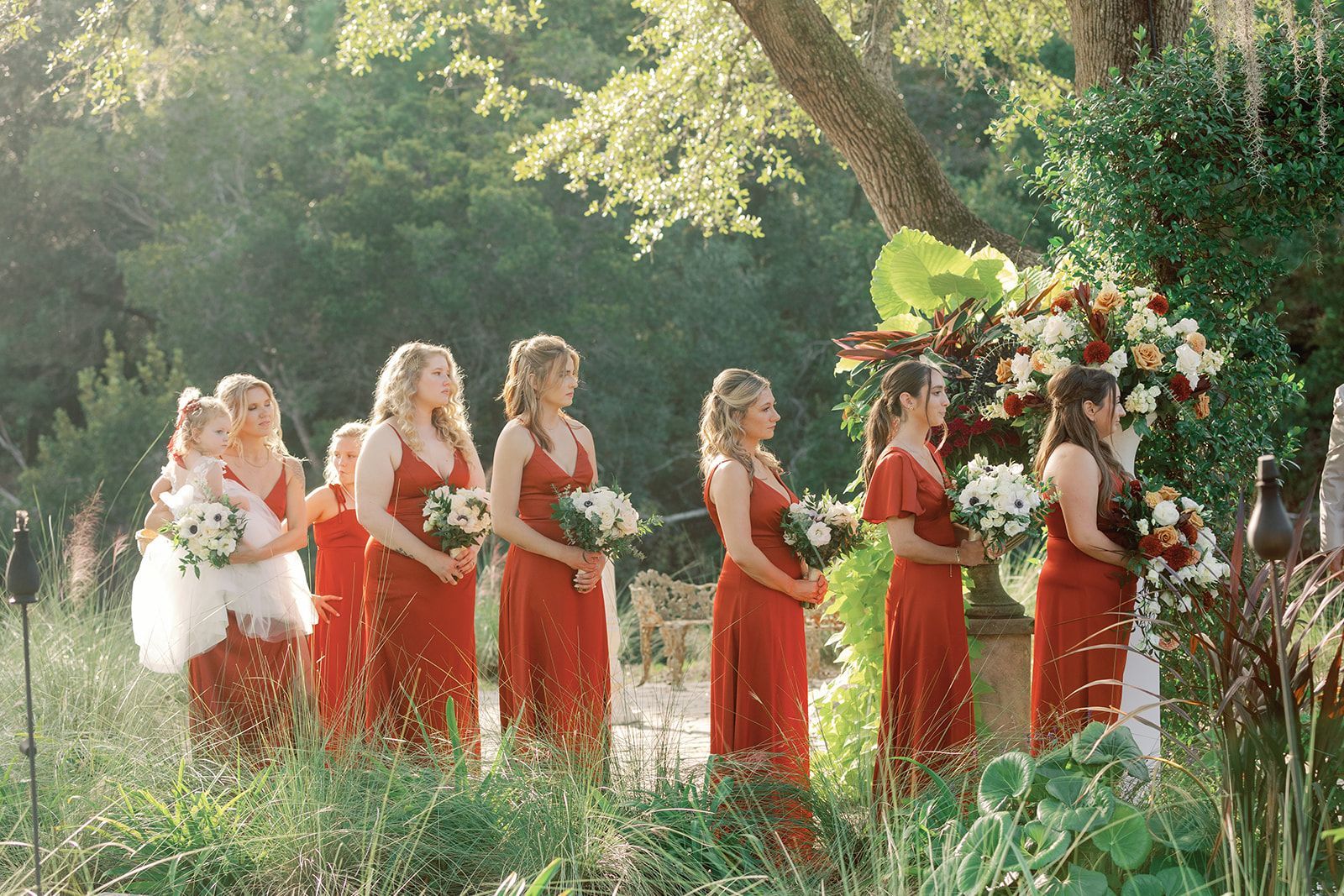 Bridesmaids in red dresses walk through a sunlit garden holding bouquets and a flower arch