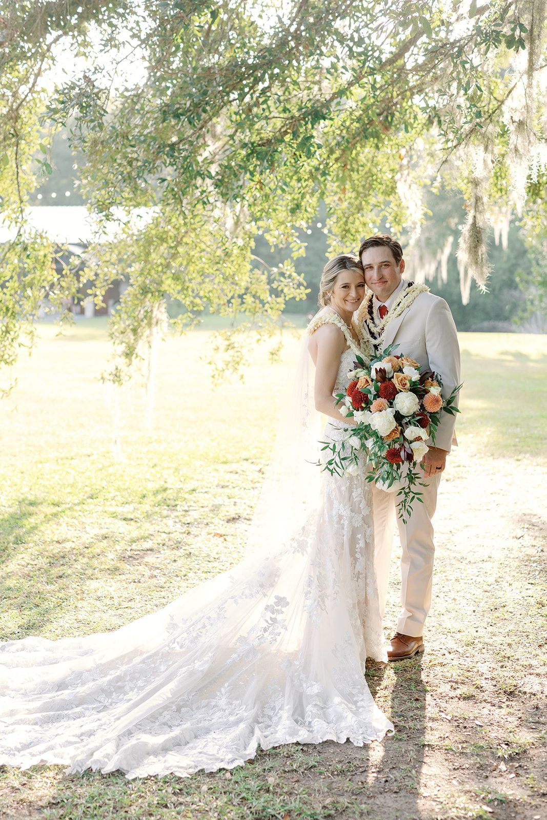Bride and groom embracing outdoors beneath a tree, with a long white gown and colorful bouquet.