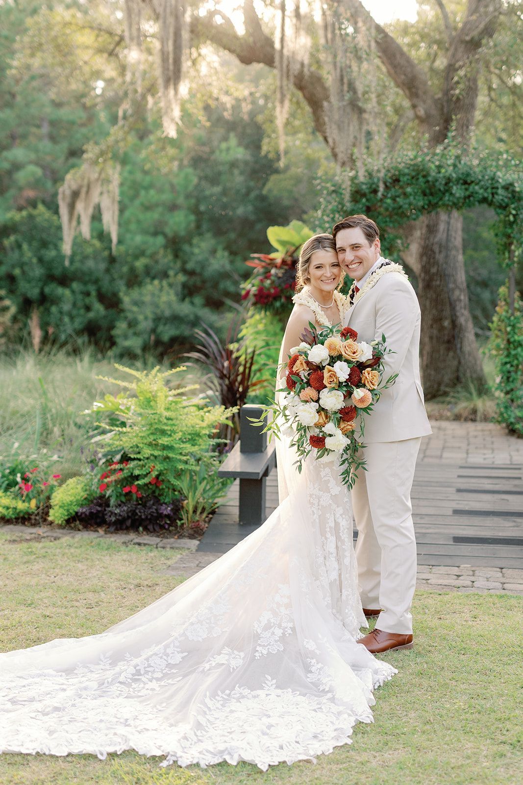 Wedding couple posing outdoors in a garden, holding a bouquet with a long lace gown and beige suit
